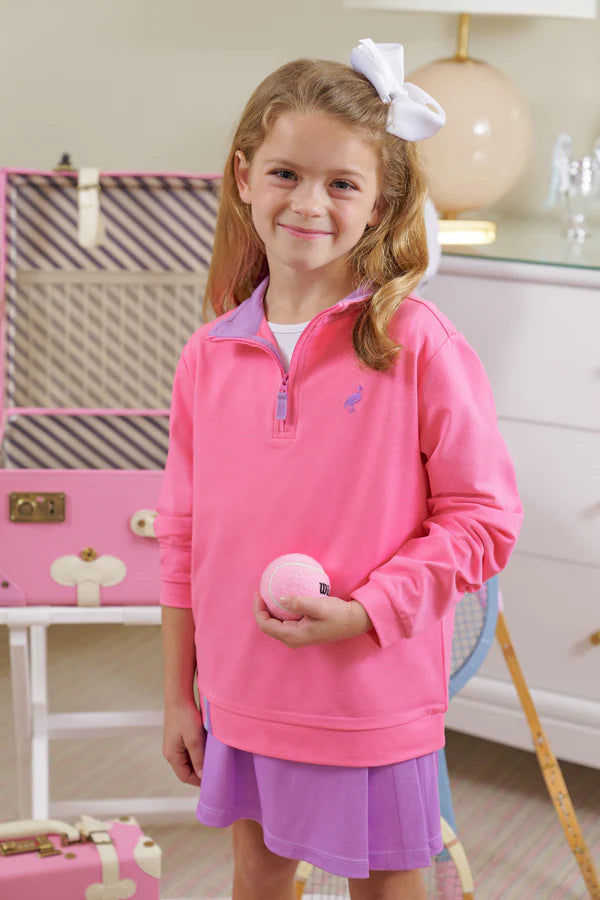 A young girl in The Beaufort Bonnet Company’s Girls' Prepletic™ Hayword Half Zip and a purple skirt holds a tennis ball indoors, with a racket and pink suitcase visible in the background.