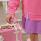 A child in a pink Girls' Prepletic™ Hayword Half Zip by The Beaufort Bonnet Company holds a small pink ball, with a pink suitcase, white shoes, and a white bottle in the background, creating a cozy and playful look.