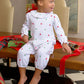 A young child in The Beaufort Bonnet Company's Baby Girls' Ramona Ruffle Romper with Christmas tree prints sits barefoot on a table, surrounded by gifts and holiday wrapping supplies.