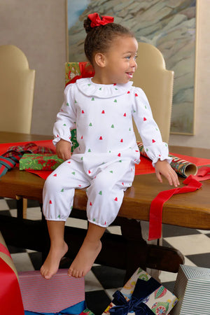 A young child in The Beaufort Bonnet Company's Baby Girls' Ramona Ruffle Romper with Christmas tree prints sits barefoot on a table, surrounded by gifts and holiday wrapping supplies.