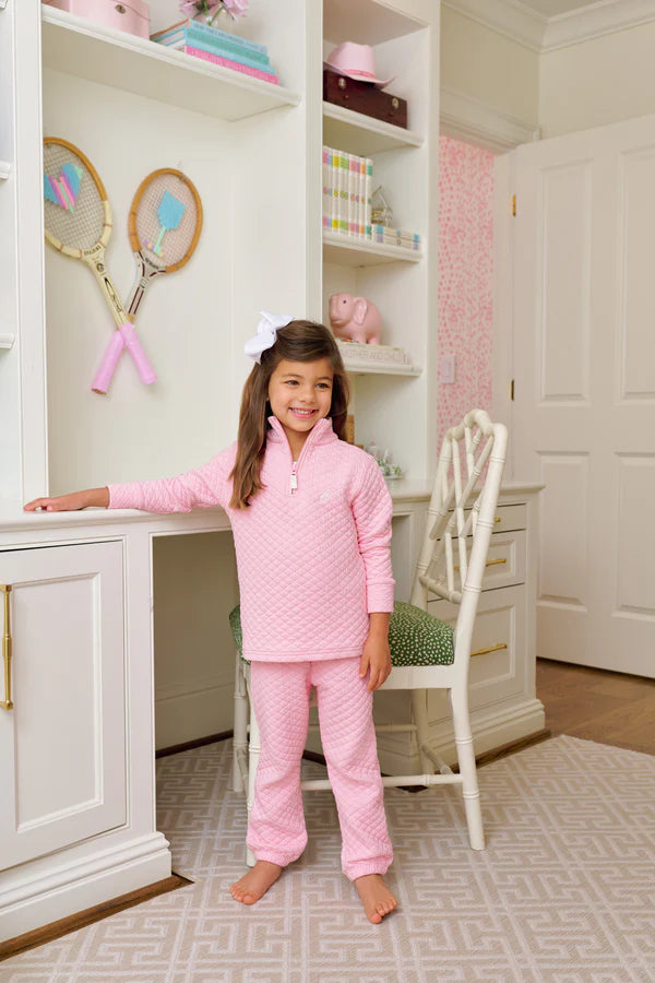 A young girl in Beaufort Bonnet Company Quilted Gates Sweeney Sweatpants stands in a white, neatly organized bedroom with a desk, chair, and tennis rackets on the wall.