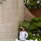 A young girl enjoys a charming picnic on the grass near a stone building, surrounded by pastel pillows, a stuffed bear, flowers, and books. She wears the Girls' Pearl Prep Skirt from The Beaufort Bonnet Company.