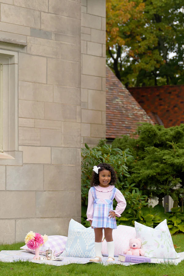 A young girl enjoys a charming picnic on the grass near a stone building, surrounded by pastel pillows, a stuffed bear, flowers, and books. She wears the Girls' Pearl Prep Skirt from The Beaufort Bonnet Company.