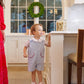 A young child wearing The Beaufort Bonnet Company's Baby Boys' Prescott Pocket Jon Jon stands on books to reach the kitchen counter, with an adult nearby, a jar of cookies on the counter, and a wreath in the window behind them.