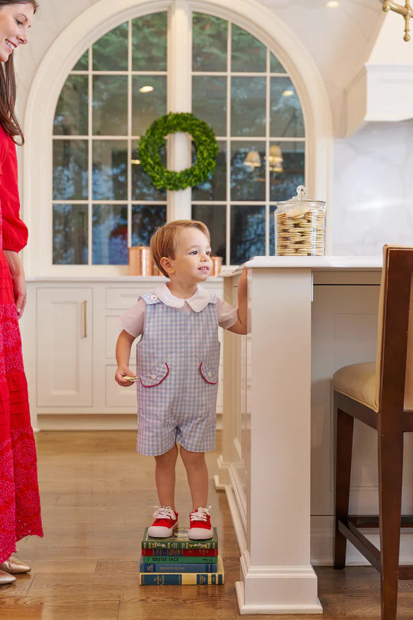 A young child wearing The Beaufort Bonnet Company's Baby Boys' Prescott Pocket Jon Jon stands on books to reach the kitchen counter, with an adult nearby, a jar of cookies on the counter, and a wreath in the window behind them.