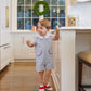 A young child in red shoes and The Beaufort Bonnet Company's Baby Boys' Prescott Pocket Jon Jon stands on stacked books in a kitchen, holding a cookie and glancing sideways. A wreath adorns the large window behind him.