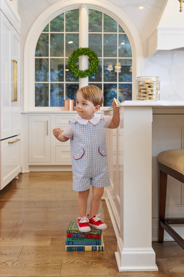A young child in red shoes and The Beaufort Bonnet Company's Baby Boys' Prescott Pocket Jon Jon stands on stacked books in a kitchen, holding a cookie and glancing sideways. A wreath adorns the large window behind him.