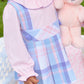 A young child wears The Beaufort Bonnet Company's Girls' Long Sleeve Ramona Ruffle Shirt and a blue plaid dress, holding a light pink teddy bear outdoors, ready for back to school.