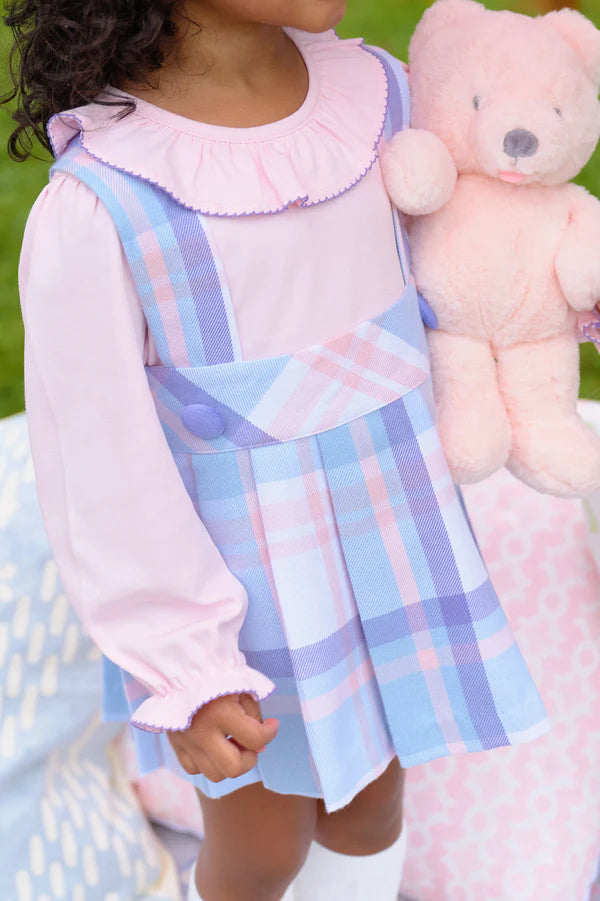 A young child wears The Beaufort Bonnet Company's Girls' Long Sleeve Ramona Ruffle Shirt and a blue plaid dress, holding a light pink teddy bear outdoors, ready for back to school.