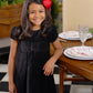 A smiling young girl with long brown hair and a red bow wears The Beaufort Bonnet Company’s Girls' Velveteen Darcy Dress as she stands beside a set dining table on a black-and-white checkered floor.