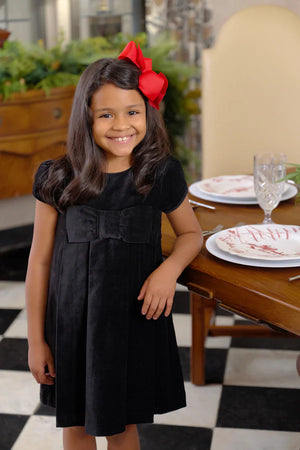A smiling young girl with long brown hair and a red bow wears The Beaufort Bonnet Company’s Girls' Velveteen Darcy Dress as she stands beside a set dining table on a black-and-white checkered floor.