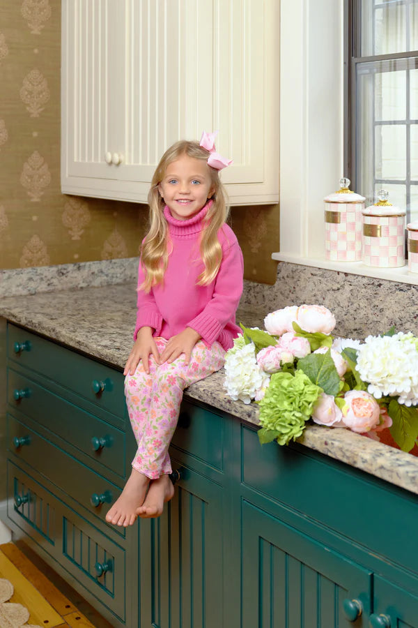 A young girl in a pink top and The Beaufort Bonnet Company Girls' Mitzy Sue Slacks sits barefoot on a kitchen counter next to flowers and decorative jars.