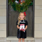 A young girl in The Beaufort Bonnet Company's Baby Girls' and Girls' Velveteen Dilly Dress stands before a door adorned with a holiday wreath, holding a wrapped gift—an ideal addition to her festive wardrobe.
