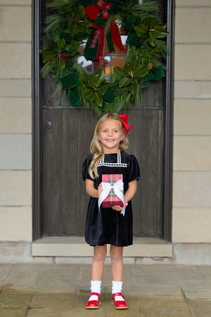 A young girl in The Beaufort Bonnet Company's Baby Girls' and Girls' Velveteen Dilly Dress stands before a door adorned with a holiday wreath, holding a wrapped gift—an ideal addition to her festive wardrobe.