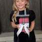 A young girl with blonde hair and a red bow wears The Beaufort Bonnet Company’s Baby Girls' and Girls' Velveteen Dilly Dress while holding a plaid gift box—an adorable pick for holiday wardrobes.