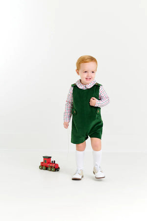 A young child in green overalls and The Beaufort Bonnet Company Baby Boys' Peter Pan Collar Shirt stands indoors, holding a string connected to a small red toy train on the floor.