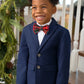 A young boy in a navy blazer, white shirt, and The Beaufort Bonnet Company's Baylor Bowtie stands smiling in front of a decorated staircase with greenery, wearing matching plaid pants.