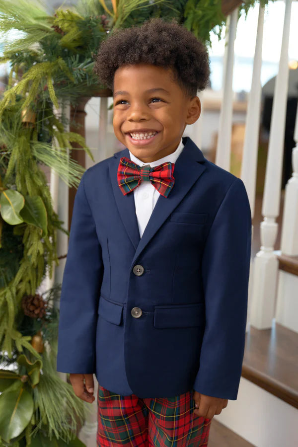 A young boy in a navy blazer, white shirt, and The Beaufort Bonnet Company's Baylor Bowtie stands smiling in front of a decorated staircase with greenery, wearing matching plaid pants.