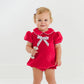 A toddler stands barefoot against a white background wearing The Beaufort Bonnet Company's Baby Girls' Banks Bow Bubble and holds a silver rattle.