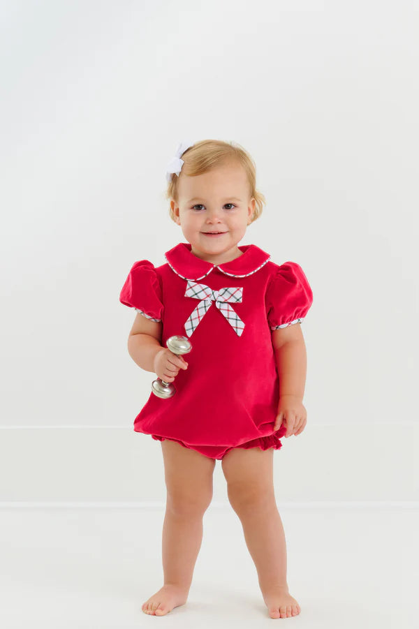 A toddler stands barefoot against a white background wearing The Beaufort Bonnet Company's Baby Girls' Banks Bow Bubble and holds a silver rattle.
