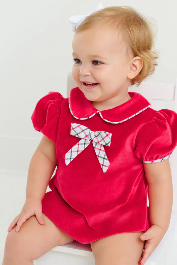 A toddler sits on a white chair, wearing The Beaufort Bonnet Company’s Baby Girls' Banks Bow Bubble with puffed sleeves and a plaid bow on the front.