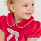 A toddler smiles and looks to the side, wearing The Beaufort Bonnet Company's Baby Girls' Banks Bow Bubble with a matching bow in her hair.