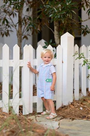 A little gent in The Beaufort Bonnet Company Baby Boys' Pima Peter Pan Collar Onesie stands by a white picket fence outdoors, surrounded by trees and plants.