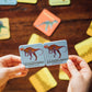 Two hands hold matching "Ceratosaurus" cards above a table with more cards from the Eeboo Dinosaurs Memory & Matching Game scattered around.