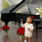 A young child in a white Baby Girls' Long Sleeve Bridget Bubble by The Beaufort Bonnet Company and red shoes stands before a black grand piano, holding a small bell. Two potted poinsettias sit near the piano.