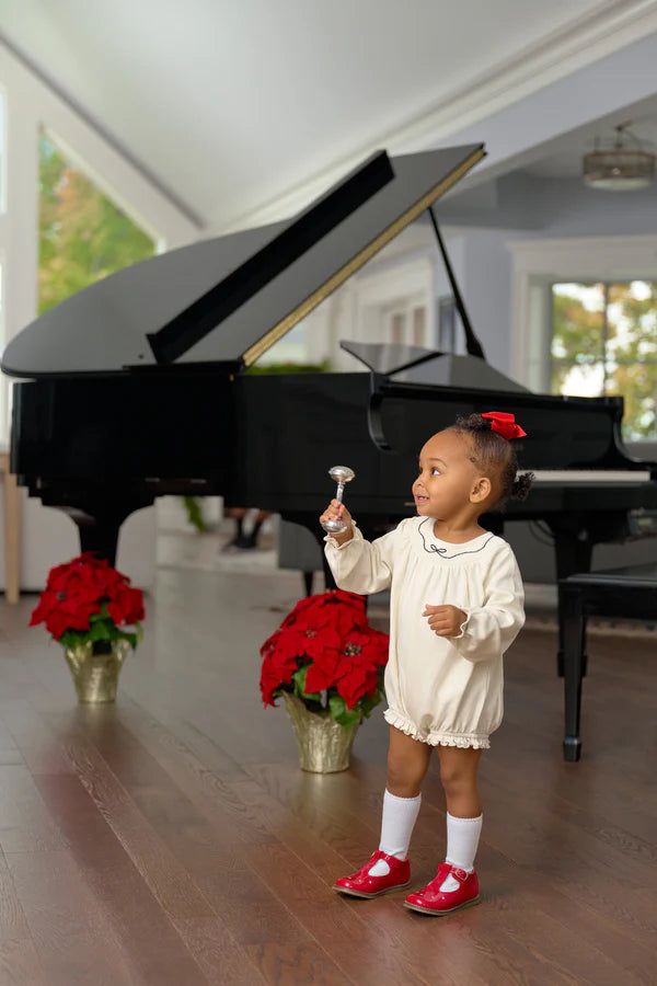 A young child in a white Baby Girls' Long Sleeve Bridget Bubble by The Beaufort Bonnet Company and red shoes stands before a black grand piano, holding a small bell. Two potted poinsettias sit near the piano.