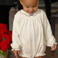 A toddler in a cream-colored Baby Girls' Long Sleeve Bridget Bubble by The Beaufort Bonnet Company stands indoors near a red poinsettia, featuring ruffled cuffs for added charm.