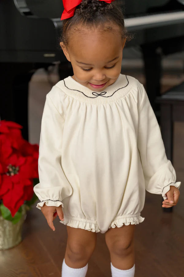 A toddler in a cream-colored Baby Girls' Long Sleeve Bridget Bubble by The Beaufort Bonnet Company stands indoors near a red poinsettia, featuring ruffled cuffs for added charm.