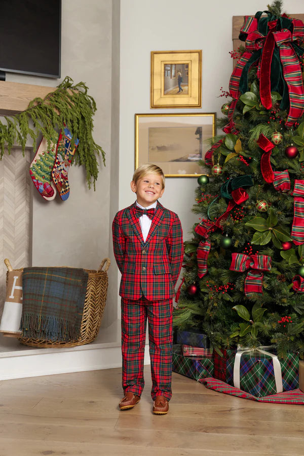 A young boy in The Beaufort Bonnet Company’s Baby Boys' and Boys' Twill Sheffield Pants stands smiling by a decorated Christmas tree with wrapped gifts below and stockings hanging on the fireplace.