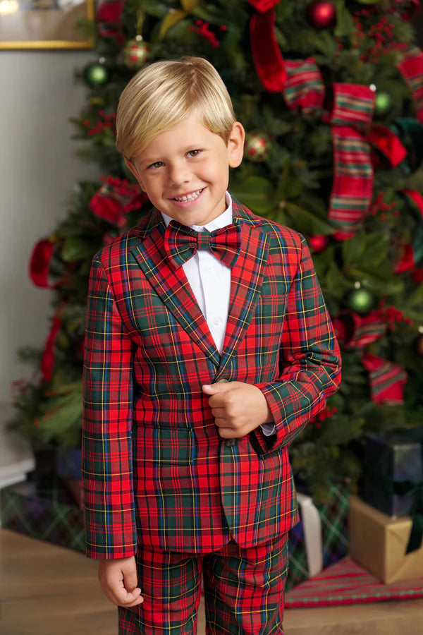A young boy wearing The Beaufort Bonnet Company’s Baylor Bowtie stands smiling in front of a decorated Christmas tree.