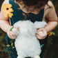 A child outdoors cuddles the Wee Ittybit Bunny by Bunnies By the Bay, holding a yellow flower in one hand and wearing a dark dress while gazing downward.
