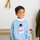 Young boy smiles indoors in the Baby Boys' and Boys' Isaacs Intarsia Sweater by The Beaufort Bonnet Company in Beale Street Blue, paired with navy pants and a white turtleneck. Snowshoes and a coat are seen in the background.