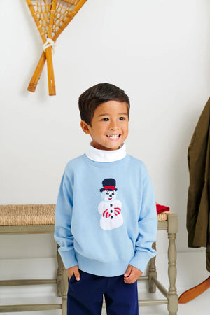Young boy smiles indoors in the Baby Boys' and Boys' Isaacs Intarsia Sweater by The Beaufort Bonnet Company in Beale Street Blue, paired with navy pants and a white turtleneck. Snowshoes and a coat are seen in the background.