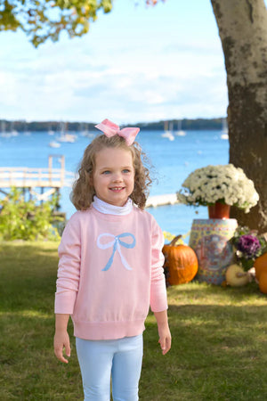A young girl wears The Beaufort Bonnet Company's Baby Girls' and Girls' Isabelle's Intarsia Sweater while standing outdoors near pumpkins and flowers, with a lake and boats in the background.