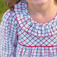 A young girl smiles outdoors in The Beaufort Bonnet Company’s Girls' Long Sleeve Ramona Ruffle Dress, showcasing a white base with red and green Old Town Tattersall plaid and a sweet ruffled collar.