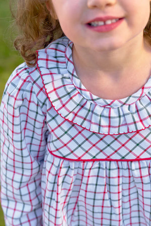 A young girl smiles outdoors in The Beaufort Bonnet Company’s Girls' Long Sleeve Ramona Ruffle Dress, showcasing a white base with red and green Old Town Tattersall plaid and a sweet ruffled collar.