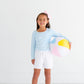 A little girl holding The Beaufort Bonnet Company Shipley Shorts, a summertime favorite, in front of a white wall.