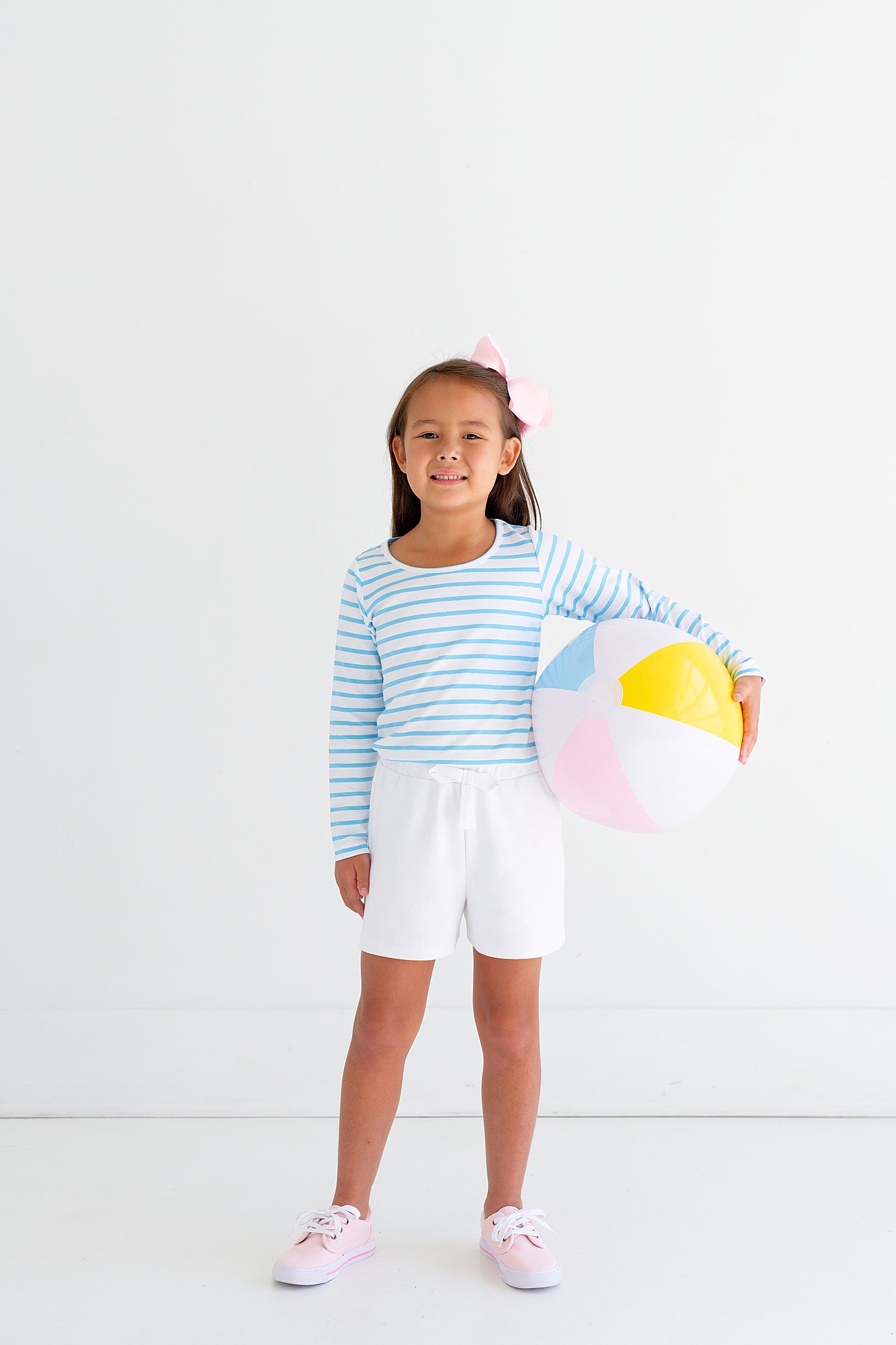 A little girl holding The Beaufort Bonnet Company Shipley Shorts, a summertime favorite, in front of a white wall.