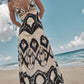 A woman in the Johanna Ortiz Zea Palm Maxi Dress, a black and white cotton poplin maxi with adjustable straps, stands barefoot on a sandy beach with the ocean and cloudy sky behind her.