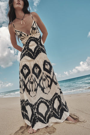 A woman in the Johanna Ortiz Zea Palm Maxi Dress, a black and white cotton poplin maxi with adjustable straps, stands barefoot on a sandy beach with the ocean and cloudy sky behind her.