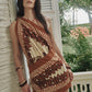 A woman stands by a white wall and railing, wearing the Johanna Ortiz Sun Traveller Mini Dress in brown and cream patterns, with palm trees and greenery in the background.