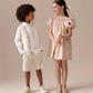 A boy in a cream vest and shorts stands beside a girl in the Bonpoint Girls' Arlene Dress, smocked and hand-embroidered in Liberty fabric, holding a flower. Both children smile in a neutral studio setting.