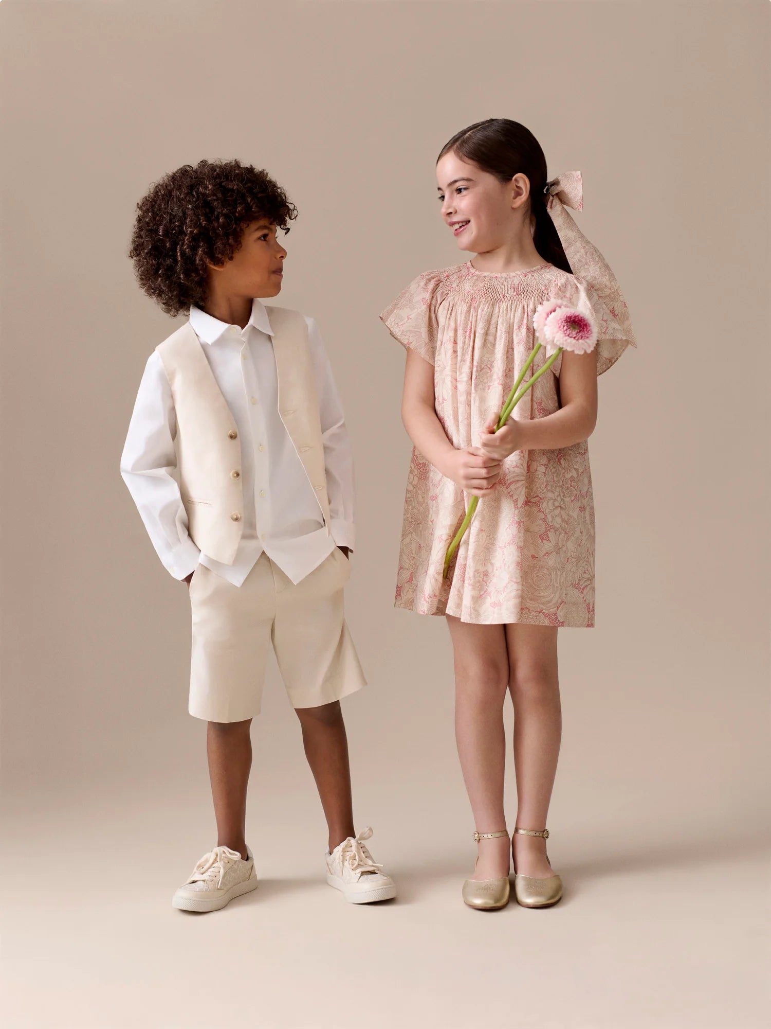 A boy in a cream vest and shorts stands beside a girl in the Bonpoint Girls' Arlene Dress, smocked and hand-embroidered in Liberty fabric, holding a flower. Both children smile in a neutral studio setting.