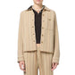 A person wearing the Dylan Stripe Shirt by Guest in Residence, featuring beige and cream stripes, paired with matching pants over a black ribbed top and a beaded necklace, shown from shoulders to thighs.