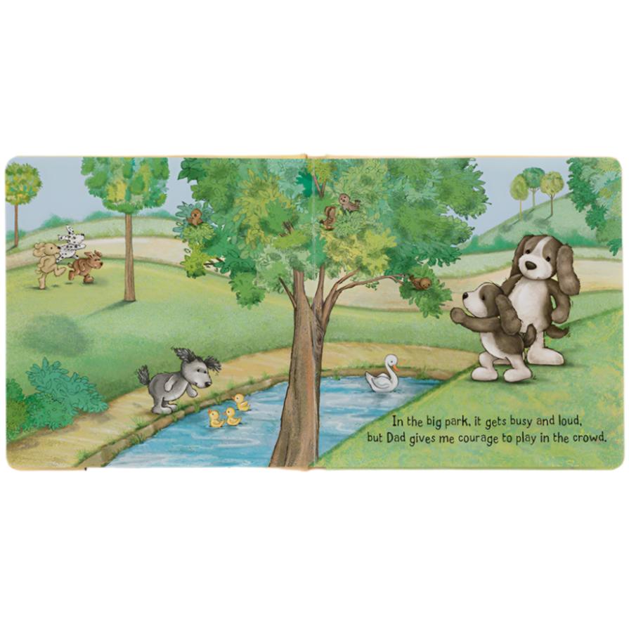 Illustrated children's book page depicting dogs in a park. Some dogs are running and playing, while one dog stands by a tree with its parent. A pond with ducklings and a swan is in the foreground. This charming scene from our "Jellycat My Dad & Me Book" captures the joy of simple adventures with daddy.