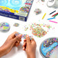 Colorful Thinking Putty is being stretched by hands on a table surrounded by craft supplies, putty containers, and small beads. A box labeled "Crazy Aarons Hide Inside Design Studio Kit" from Crazy Aarons, along with colorful pens, sets up the perfect scene for sensory integration fun.